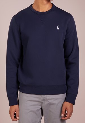 Mężczyzna ubrany w granatowy, długorękawowy sweatshirt z okrągłym dekoltem i małym białym logo gracza polo na lewej piersi oraz jasnoszare spodnie.