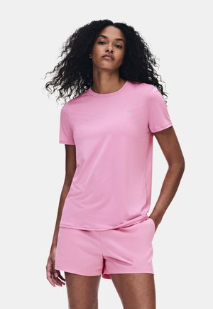 Donna con capelli ricci che indossa una maglietta rosa chiaro tinta unita a maniche corte e pantaloncini abbinati su uno sfondo chiaro e uniforme.