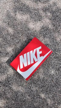 Caixa Nike vermelha com um logótipo branco proeminente e design de swoosh. Textura suave, forma retangular, posicionada sobre uma superfície cinza texturizada.