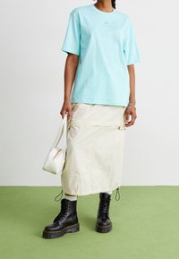 Ljusblå bomulls-t-shirt, krämfärgad cargoskirt med fickor, vit handväska och svarta snörkängor med tjock sula på grön golv.