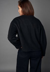 Schwarze, cropped Jacke aus einem strukturierten Stoff, mit weiten Ärmeln und einem schlichten Design. Über dunklen Hosen getragen, von hinten betrachtet.