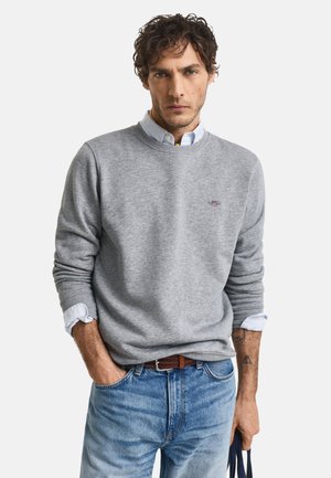 Grauer Pullover mit Rundhalsausschnitt, weichem Stoff und gesticktem Logo auf der Brust, kombiniert mit hellblauen Jeans und einem braunen Gürtel.