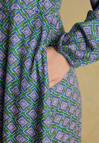 Šaty s dlhým rukávom s geometrickým vzorom v modrej, zelenej a žltej farbe a s vreckom. Látka má hladkú textúru a jemný lesk.
