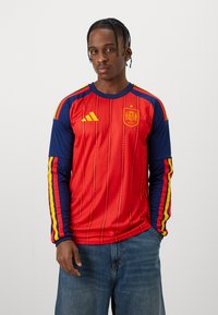 Man med flätat hår som bär en långärmad röd och marinblå spansk fotbollströja med gula ränder och blå jeans, stående med handen i fickan.