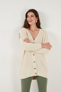 Beige neulecardigan, jossa on V-pääntie, pitkät hihat ja nappikiinnitys. Varustettu ribbireunuksilla hihansuissa ja helmassa, sekä rento malli.