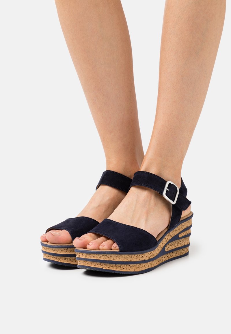 Pieds portant des sandales compensées en daim bleu marine avec des semelles compensées en liège et des brides de cheville avec des boucles argentées.