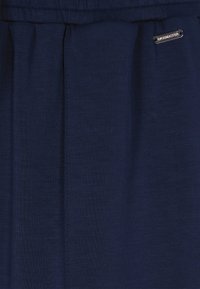 Navyblauwe stof met een zachte textuur, voorzien van plooien en een metalen logo-accent. De tailleband is elastisch, wat zorgt voor een comfortabele pasvorm.