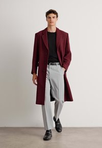 Palton bordeaux din lână cu nasturi mari, asortat cu pantaloni gri, cămașă neagră și pantofi negri. Textură netedă și croială ajustată.