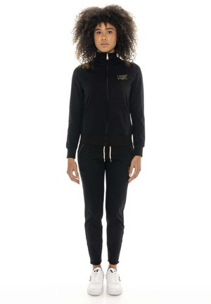 Une femme porte une veste noire zippée assortie à un pantalon de jogging, avec des baskets blanches, sur un fond blanc uni.