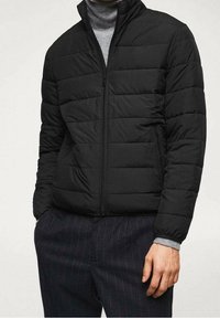 Veste rembourrée noire avec col montant, présentant des sections matelassées horizontales et une fermeture éclair à l'avant. Portée sur un col roulé gris.