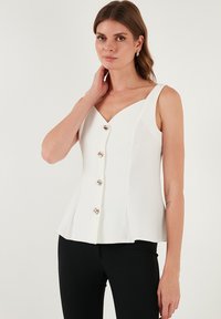 Fehér, ujj nélküli felső V-kivágással, fodros szegéllyel és hat ezüstgombbal az elején. Simasága miatt mintázat nélküli anyag.