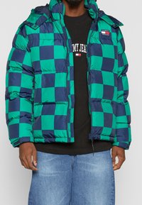 Green and navy checkered puffer jacket with a hood, zip closure, and elasticized cuffs, worn over a black sweatshirt.