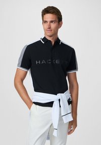 Camisa polo negra con acentos grises y ribete blanco, con el logo "HACKETT" en el pecho. Combinada con pantalones blancos y un suéter azul claro atado a la cintura.