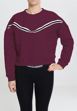 Bordeauxfarbener Pullover mit weißen und schwarzen diagonalen Streifen über der Brust, cropped Design, gerippte Bündchen und Saum, kombiniert mit schwarzen Leggings.