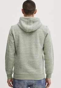 Sweat-shirt à capuche en matière polaire vert clair, avec une finition texturée, des poignets et un ourlet côtelés. Détails de couture visibles au dos.