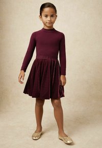 Bordová šaty s dlhými rukávmi a plisovanou zamatovou sukňou, skombinované so zlatými baletkami. Hladká textúra, priliehavý korzet a jemný detail na páse.