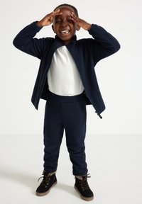 Navy blue fleece jacket with high collar and zip, paired with navy pants and black boots with faux fur lining. Child shows joyful expression.