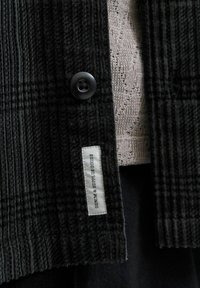 Czarna i szara kurtka w kratę z weluru z czarnym guzikiem, z wplecioną metką z napisem "DENIM & SUPPLY GOODS" po boku.