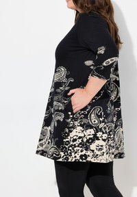 Musta tunikamekko, jossa on paisley- ja kukkakuoseja kreeminsävyssä. Kolme neljännesosaa hihasta ja sivutaskut lisäävät toiminnallisuutta ja muotoilua.