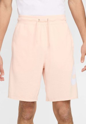 Shorts à cordon rose clair portés par une personne, avec un grand logo blanc sur le côté et une taille élastique.