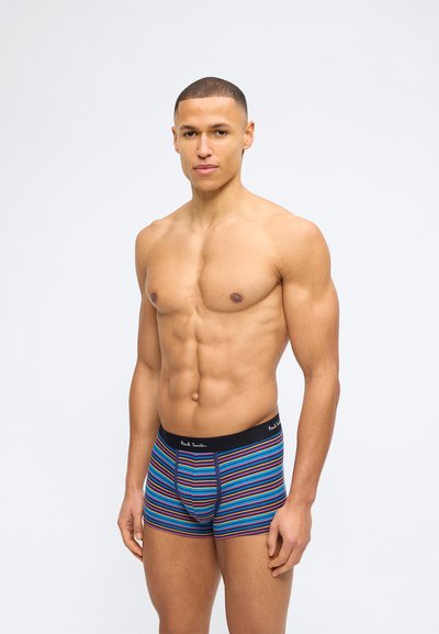 Homme torse nu debout, portant un boxer rayé multicolore avec une ceinture noire, sur un fond blanc uni.