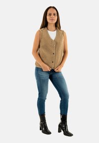 Gilet smanicato beige, con bottoni, realizzato in un materiale felpato testurizzato, indossato sopra una camicia bianca, abbinato a jeans blu e stivali neri con tacco.