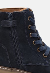 Tmavě modré semišové kotníkové boty s bočním zipem, kulatými tkaničkami, zlatými oky a kontrastními švy.