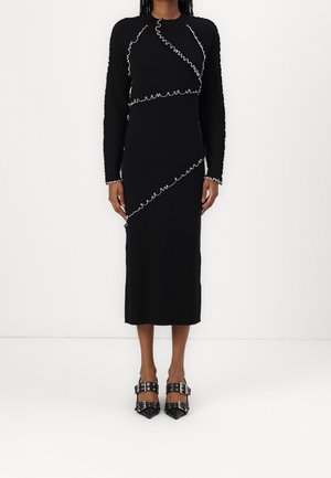 Femme portant une robe midi noire à manches longues avec des détails de bordure ondulée blanche et des chaussures noires à bouts pointus avec des brides bouclées.