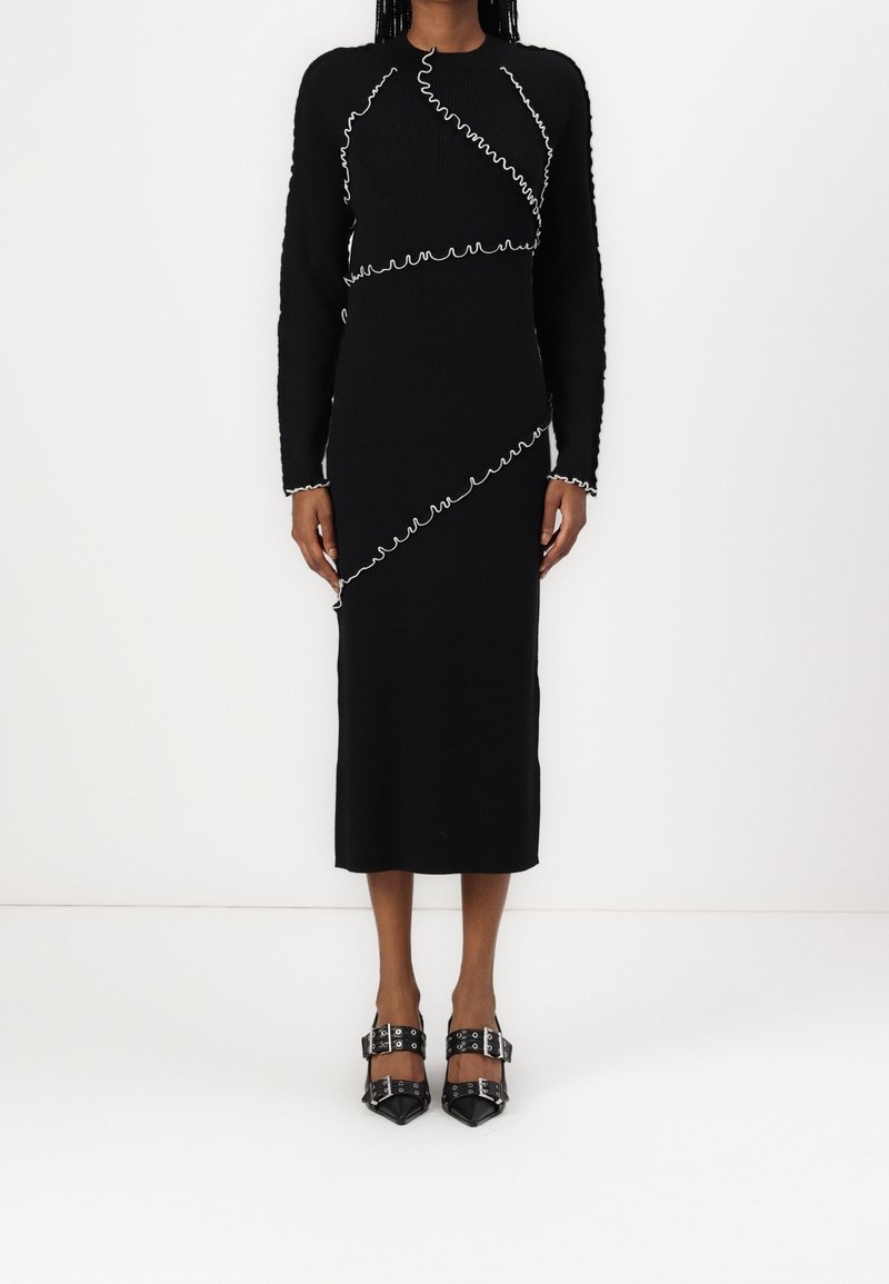 Femme portant une robe midi noire à manches longues avec des détails de bordure ondulée blanche et des chaussures noires à bouts pointus avec des brides bouclées.