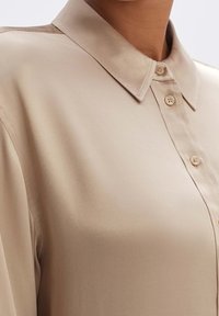 Beżowa satynowa bluzka z kołnierzykiem, z zapięciem na guziki z przodu oraz gładką fakturą; charakteryzuje się delikatnym połyskiem i zdobioną formą.