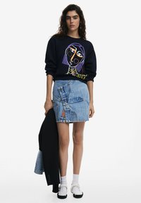 Sudadera negra con gráfico colorido, falda de mezclilla azul clara con dobladillo en ángulo y detalle de botones, combinada con calcetines blancos y zapatos negros.