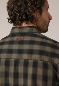 Man met krullend haar die een groen en zwart geblokt shirt draagt met een leren label op de bovenrug.