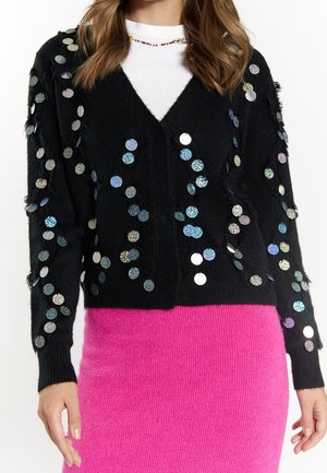 Femme portant un cardigan noir avec des sequins ronds brillants sur un haut blanc et une jupe côtelée rose vif.