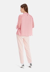 Bluse mit rosa Punkten und transparenten Ärmeln, mit geknöpftem Kragen und Knopfdetail auf der Rückseite. Kombiniert mit hellrosa Hosen und silbernen Stiefeln.