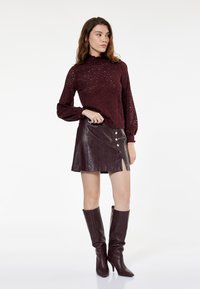 Maglione bordeaux lavorato a maglia con motivo testurizzato, collo alto e maniche a sbuffo abbinato a una minigonna in pelle marrone con dettagli a bottoni.