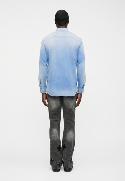 Άνδρας που στέκεται με την πλάτη, φοράει ανοιχτό μπλε τζιν πουκάμισο και γκρι τζιν με φθορές και μαύρα σταυρωτά μπαλώματα στα πίσω πόδια.
