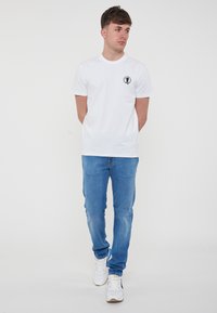 T-shirt bianco in cotone con maniche corte, dotato di un piccolo logo nero sul lato sinistro del petto. Indossato con jeans azzurri chiari e sneakers bianche.