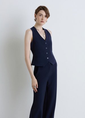 Donna con capelli castani corti che indossa un gilet smanicato blu navy abbottonato e pantaloni a palazzo coordinati, in piedi davanti a uno sfondo chiaro uniforme.