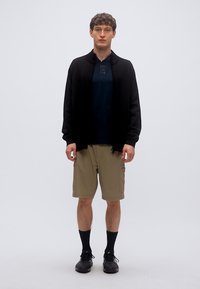 Μαύρη μακριά ζακέτα με μακριά μανίκια πάνω από μπλε σκουρόχρωμη μπλούζα, συνδυασμένη με μπεζ cargo σορτς και μαύρες κάλτσες και παπούτσια. Απλός και λειτουργικός σχεδιασμός.