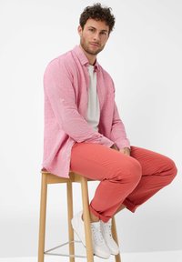 Camicia button-up rosa chiaro con una sottile texture a righe, abbinata a pantaloni corallo vivaci e sneakers bianche. Seduto su uno sgabello di legno.