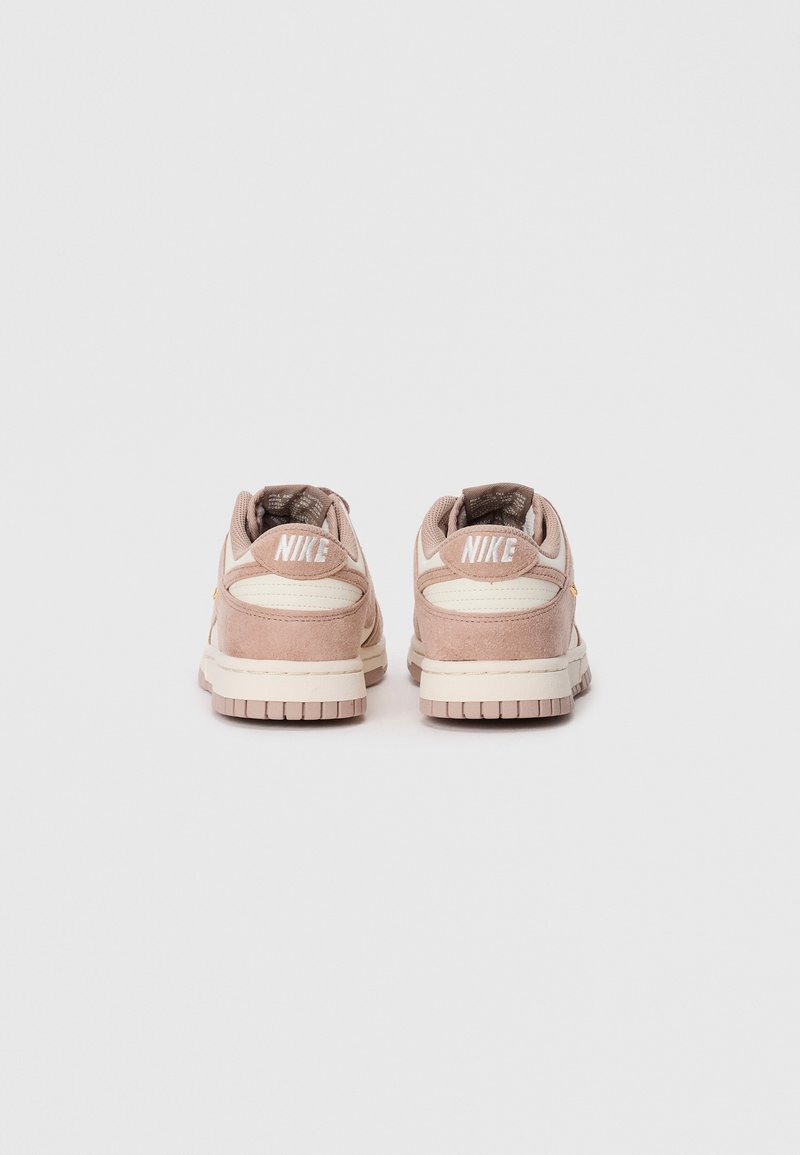 Sneaker in suede rosa con accenti bianchi e colli imbottiti. Presenta una suola piatta in gomma e il logo “Nike” sul retro.