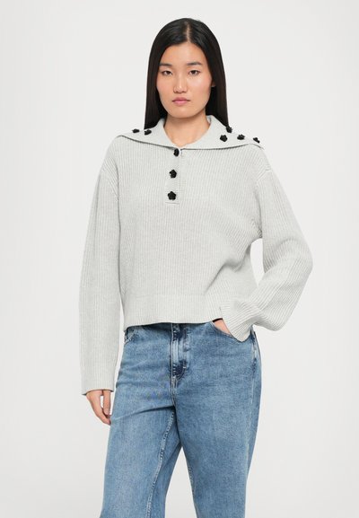 Pull en tricot gris clair avec un col, doté de détails de boutons noirs et d'un tissu texturé. Associé à un jean bleu.