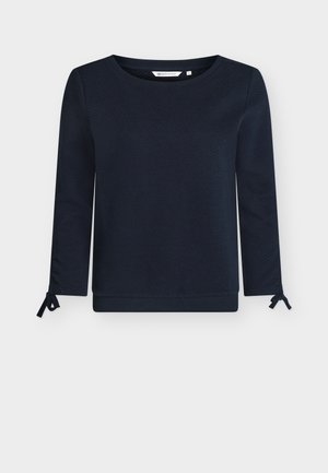 Μπλούζα μακρυμάνικη σε ναυτικό μπλε με υφή υφάσματος, στρογγυλή λαιμόκοψη και δεσίματα στα μανίκια. Ολοκληρώνεται με ελαστικό τελείωμα σε μονόχρωμο χρώμα.