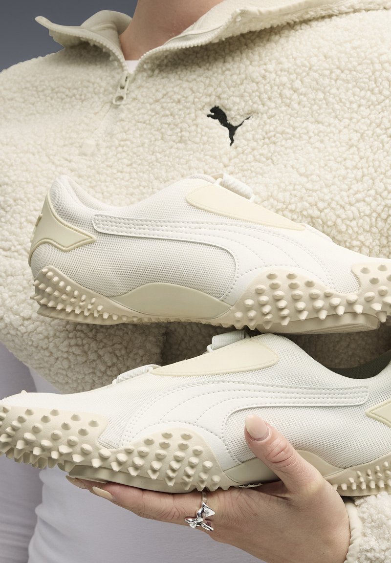 Weiße Sportschuhe bestehen aus einer Kombination aus strukturiertem Mesh und glattem Material, mit einer genoppten Sohle und minimalistischen Designelementen.