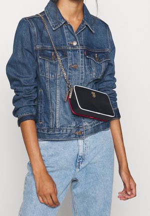 Borsa rettangolare nera e rossa con bordo bianco, tracolla a catena e logo dorato. Viene indossata a tracolla sopra una giacca di jeans.