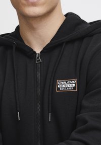 Zwarte zip-up hoodie met een logo patch op de voorkant in oranje en witte tekst, met trekkoorden en een geborstelde stofstructuur.