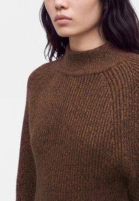 Femme aux cheveux foncés portant un pull en maille côtelée marron avec un col rond haut sur un fond uni.