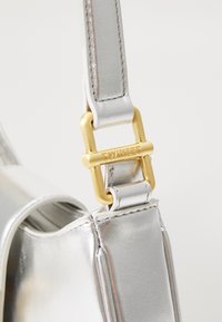 Bandoulière de sac en cuir argenté attachée à une boucle en métal doré gravée du nom de la marque "TWINSET".