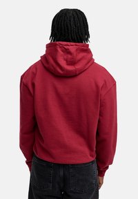 Rote, cropped Kapuzenjacke mit einer großzügigen Kapuze, aus einem Baumwoll-Mischgewebe, mit gerippten Bündchen und einem lässigen Schnitt. Kombiniert mit schwarzen, strukturierten Jeans.