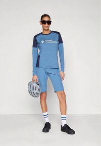 Mėlynas ilgomis rankovėmis sportinis marškinėlis su tamsiai mėlynomis detalėmis, logotipas priekyje; derantys šortai, juodi batai, akiniai nuo saulės ir pilkas šalmas rankoje.